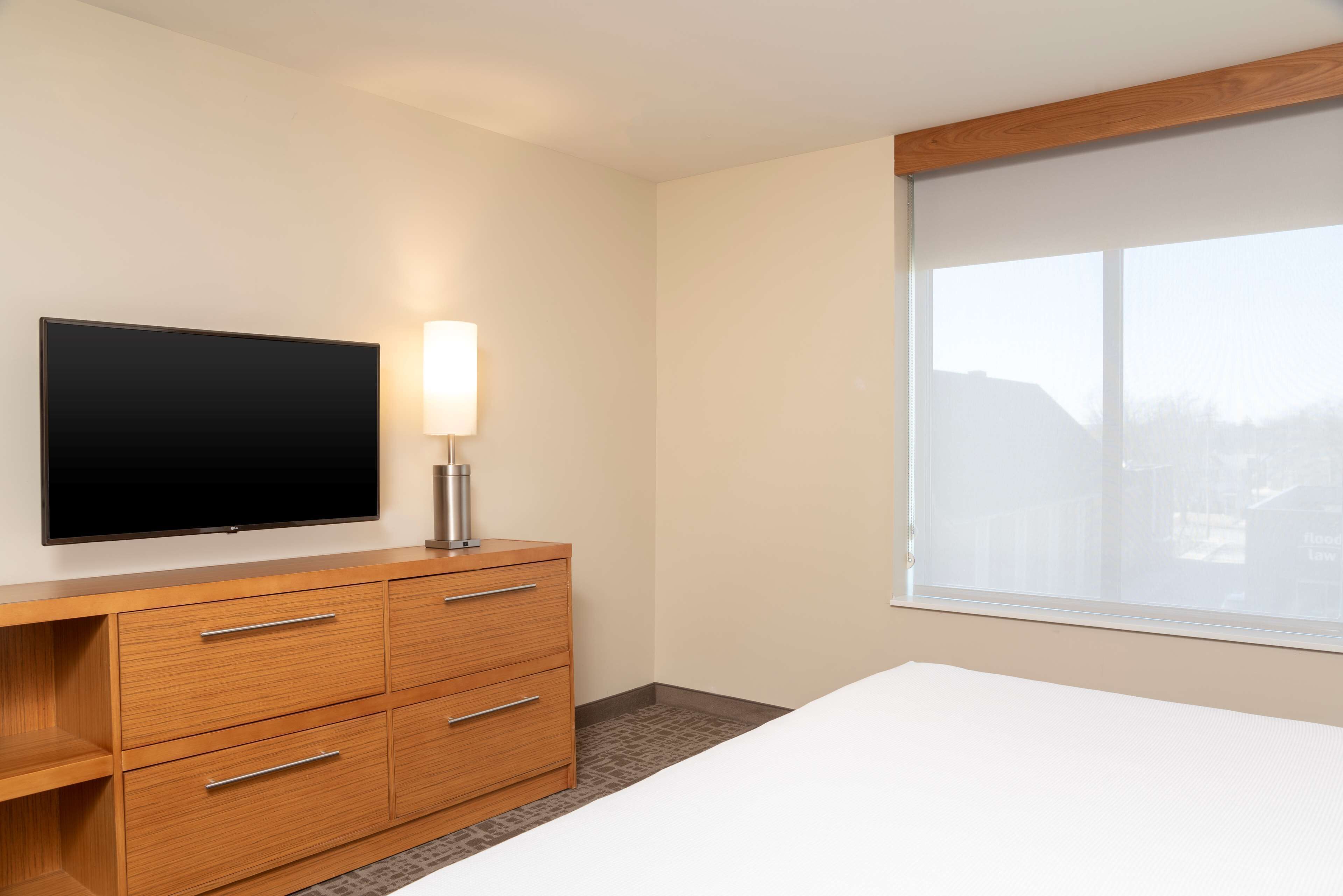 Une chambre d’hôtel avec un lit, une commode en bois dotée de tiroirs, un téléviseur à écran plat fixé au mur et une fenêtre donnant sur des immeubles.