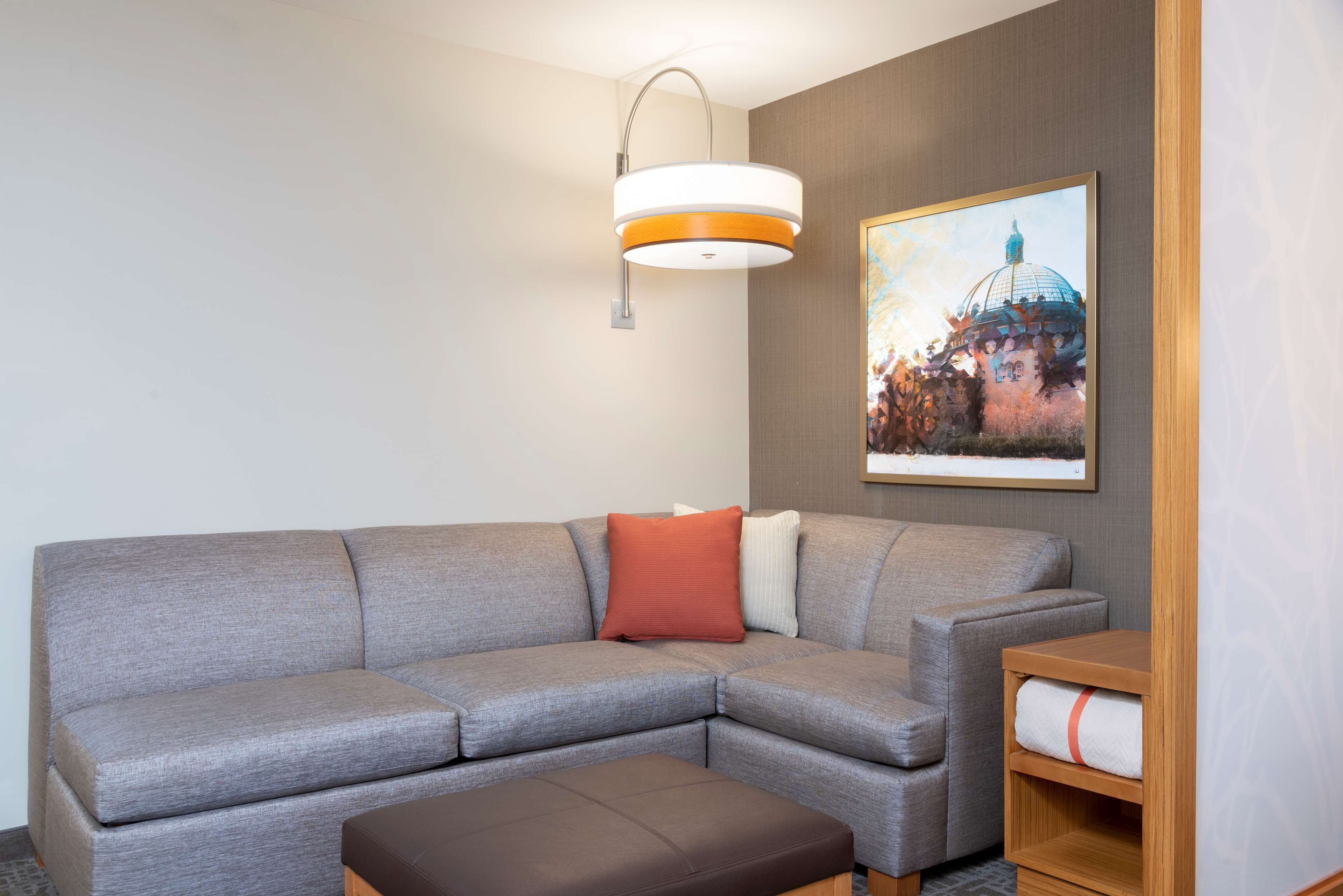 Una sala de estar moderna con un sofá seccional gris, un reposapiés marrón, una lámpara de pared y un cuadro enmarcado de un edificio.