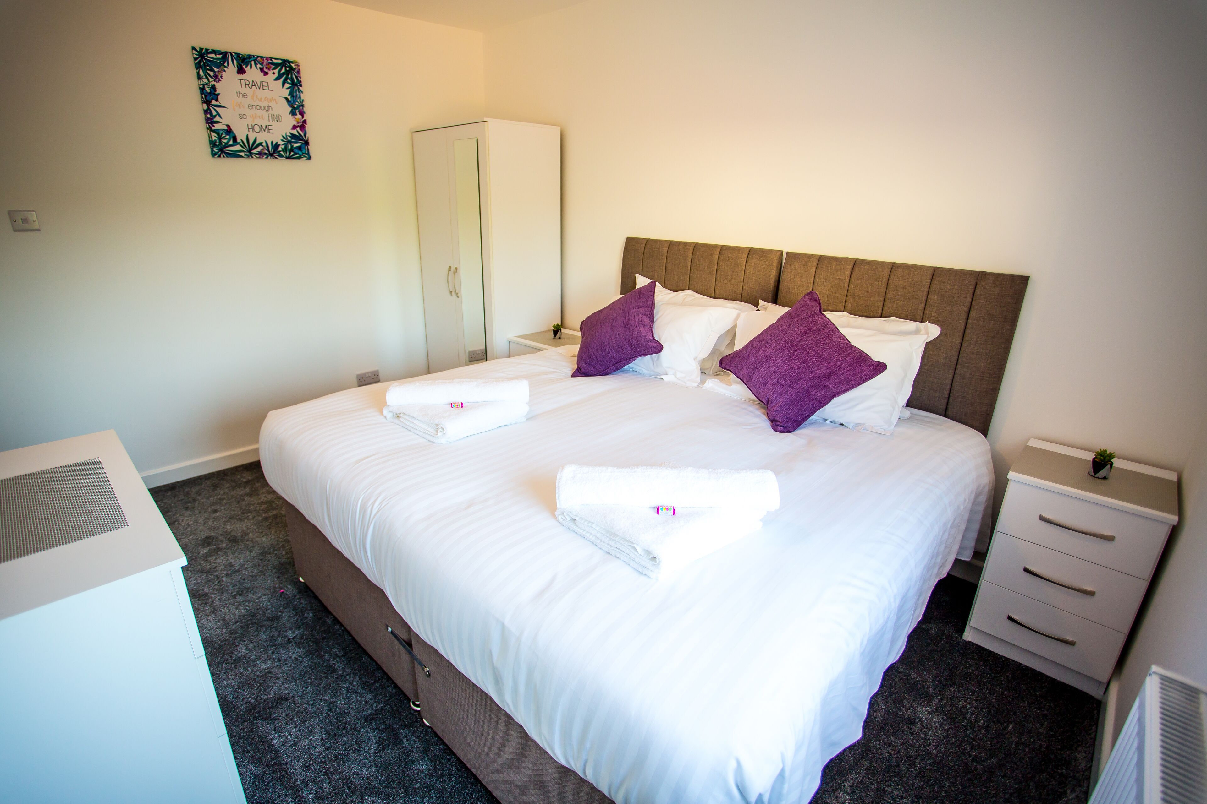 A neatly made bed with white linens and purple pillows, a white wardrobe, a small bedside table with a lamp, and a wall-mounted sign.