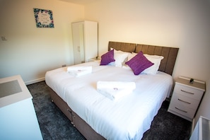 A neatly made bed with white linens and purple pillows, a white wardrobe, a small bedside table with a lamp, and a wall-mounted sign.