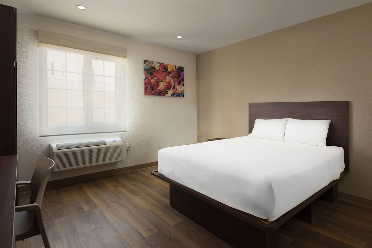 Hotelkamer met een groot bed, een bureau, een stoel, een raam met jaloezieën en een kleurrijk schilderij aan de muur.