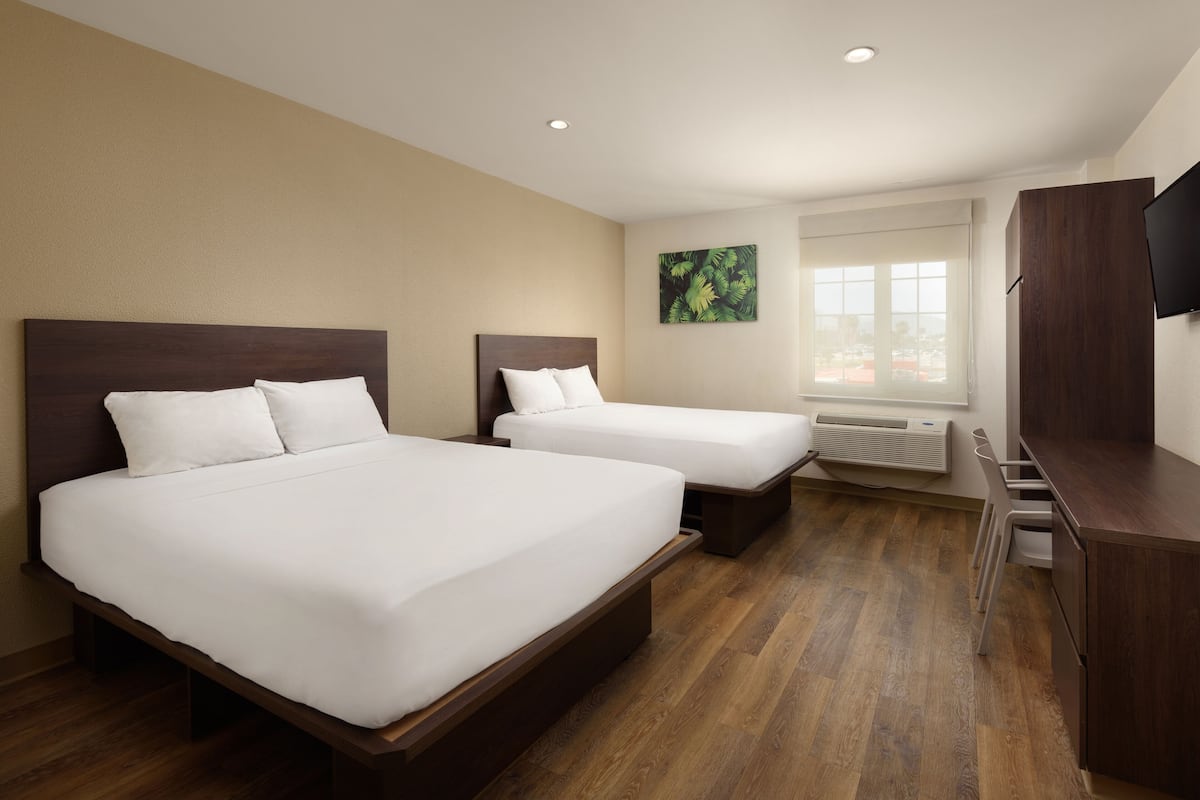 Een hotelkamer met twee bedden, een televisie, een raam met jaloezieën en een schilderij aan de muur.
