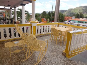 Un espace extérieur abrité avec des chaises et des tables jaunes, une rambarde blanche agrémentée d’éléments décoratifs, et une vue sur un paysage montagneux en arrière-plan.