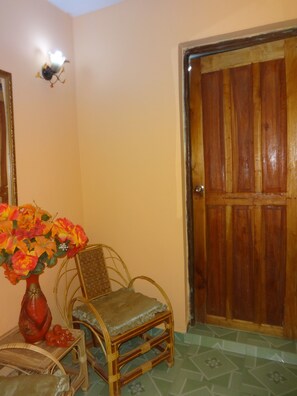 Une pièce avec une porte en bois, une chaise en osier, une petite table sur laquelle est posé un vase de fleurs et un luminaire fixé au mur.