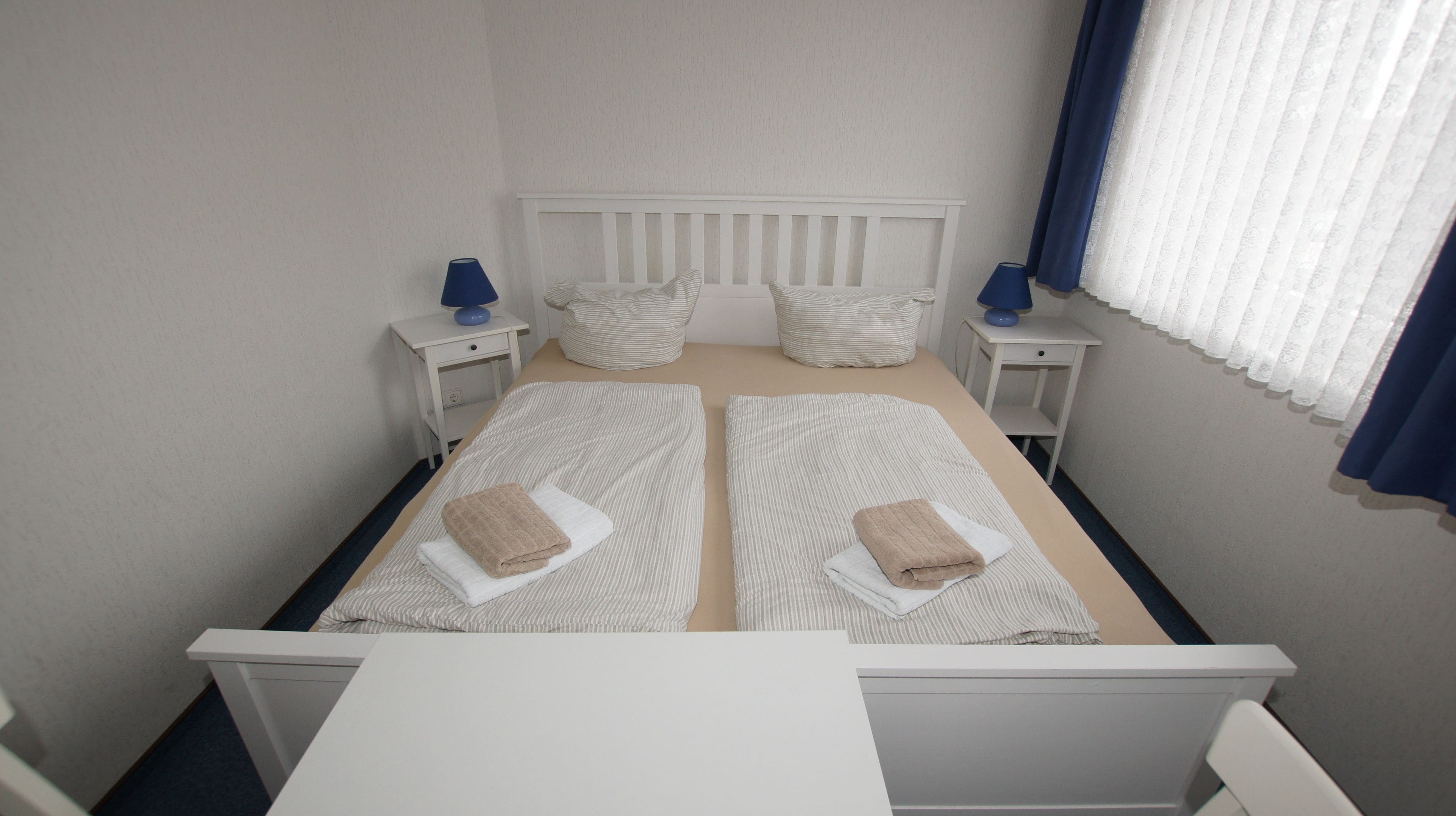 Ein Doppelbett mit zwei Kissen, ein Nachttisch mit Lampe und ein Fenster mit Vorhängen.
