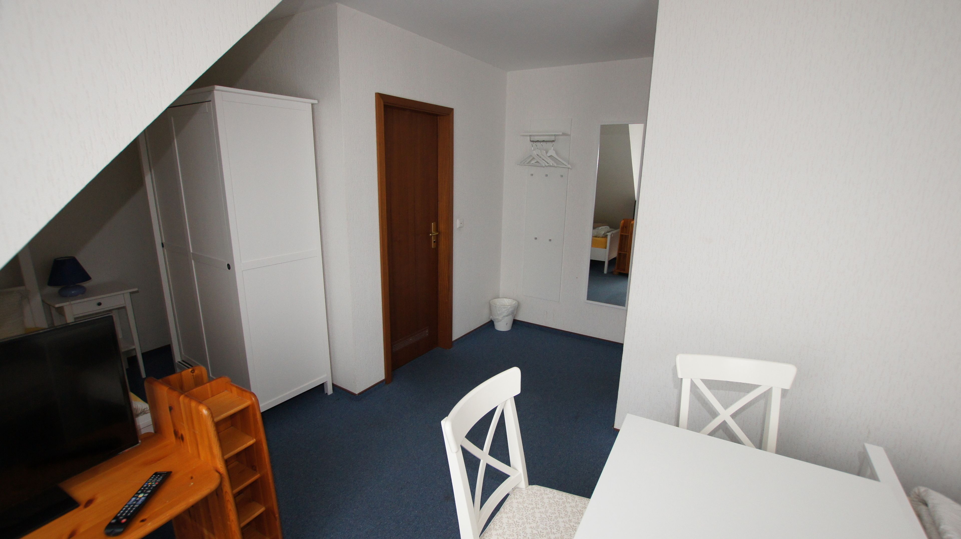 Een kleine kamer met een schuine kap, een wit bureau, een stoel, een televisie, een garderobe en een deur.