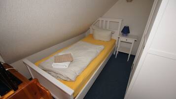 Ein kleines Zimmer mit einem Einzelbett, einem Holzbettrahmen, einer gelben Matratze und einem weißen Kopfteil. Ein Nachttisch mit Lampe und kleiner Schublade ist vorhanden.