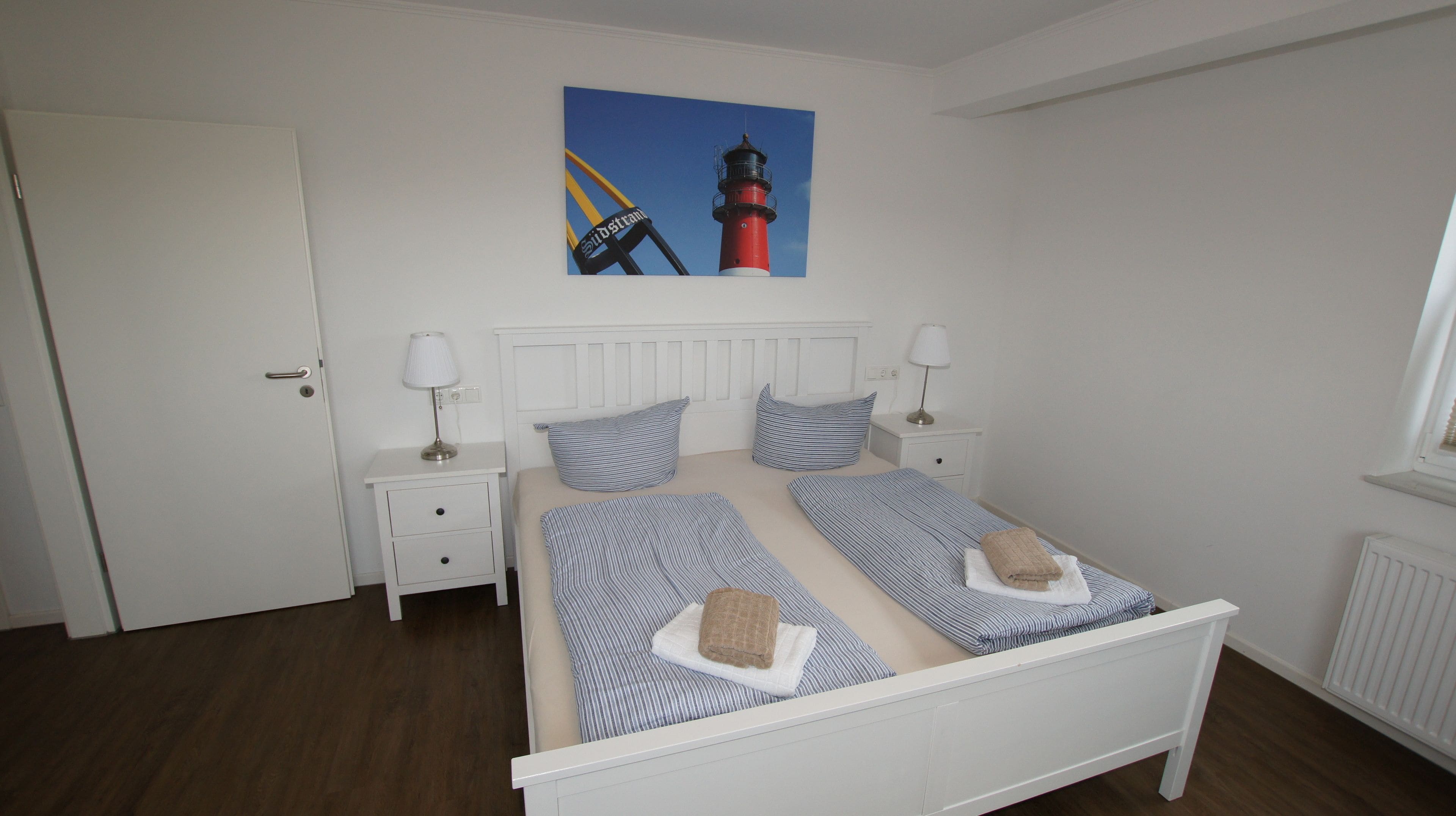 Une chambre à coucher bien rangée, avec un lit, deux tables de chevet, une commode, une fenêtre et un tableau représentant un phare.