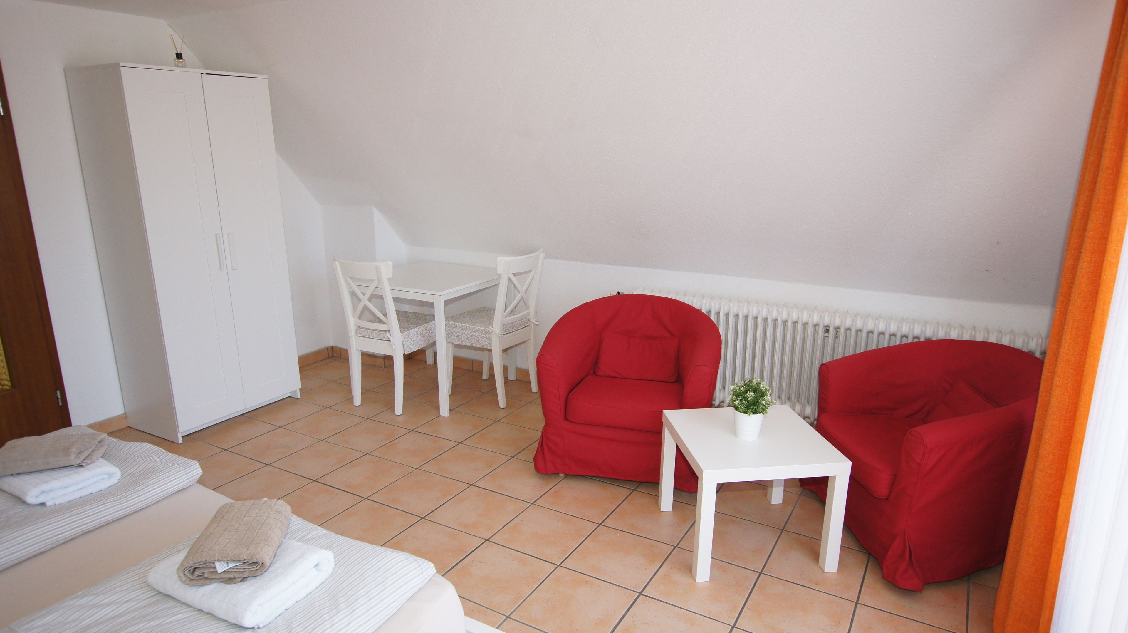 Una habitación pequeña y limpia con un armario blanco, una mesa blanca con dos sillas, un sofá rojo y una mesita blanca con una planta.