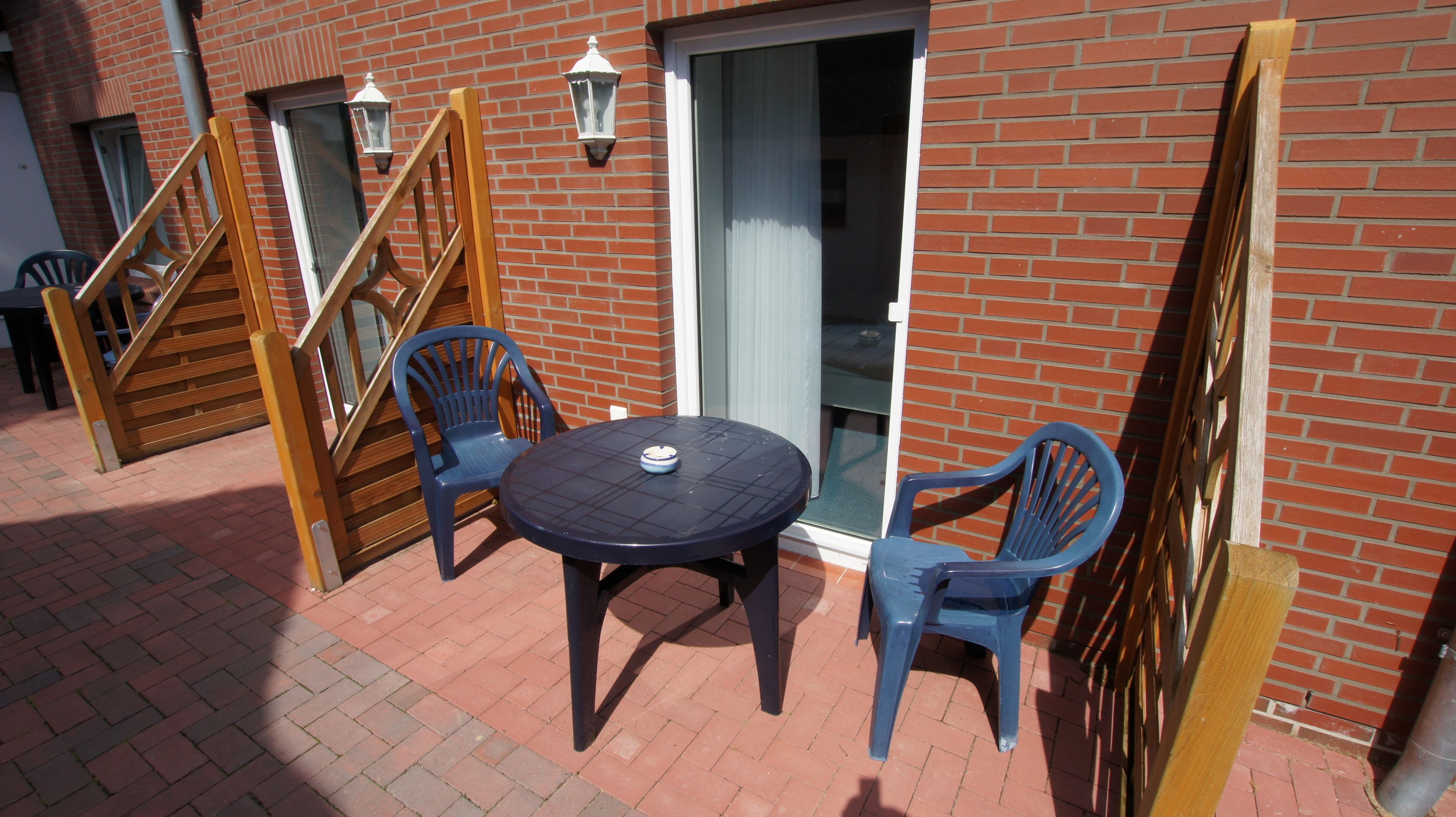 Eine Terrasse mit rundem Tisch und zwei Stühlen, eine Holztreppe und eine Backsteinmauer.