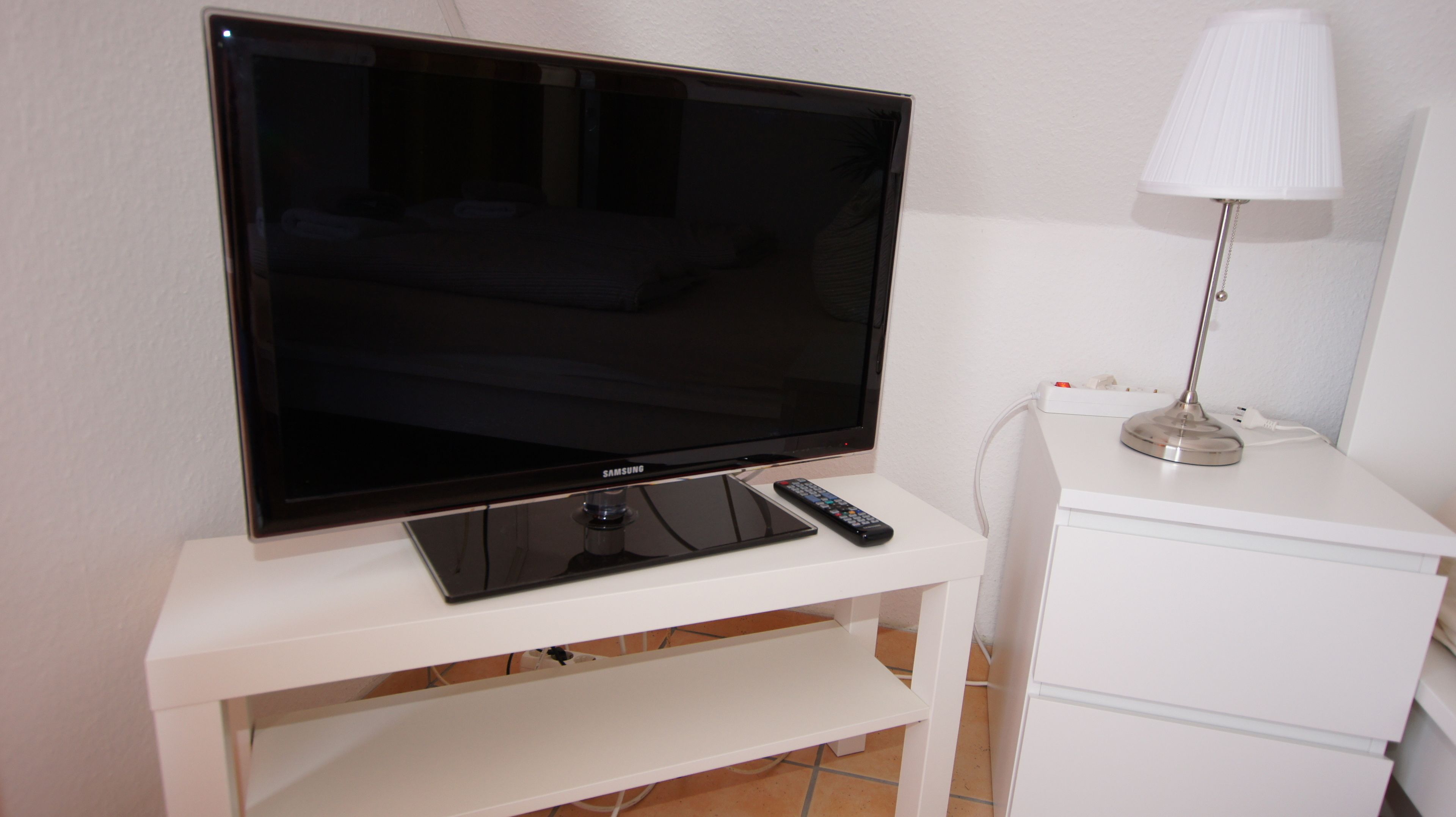 Un televisor sobre un soporte blanco con un control remoto, una lámpara y un enchufe.