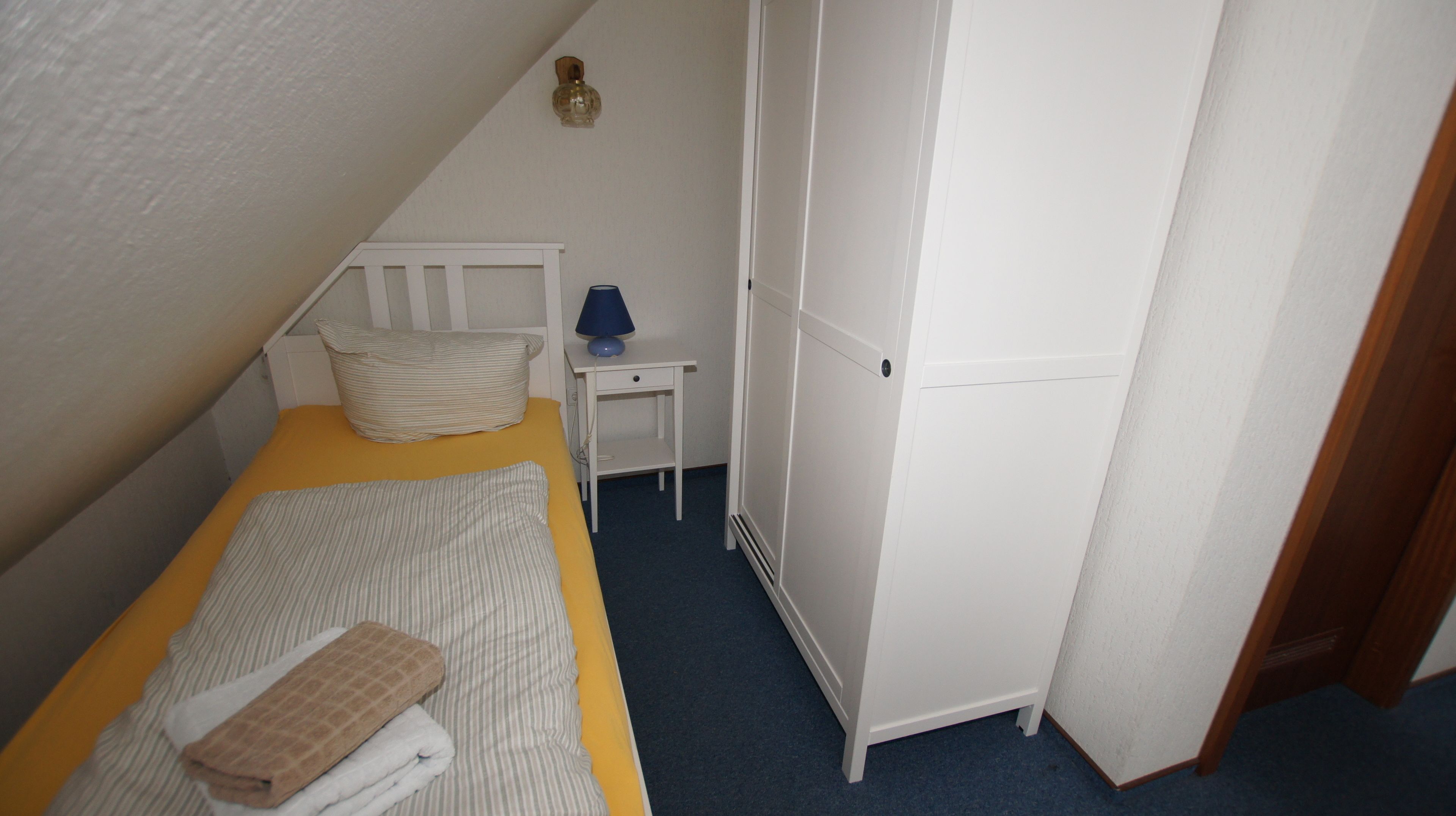 Un dormitorio pequeño y ordenado con una cama individual, una mesita de noche con lámpara y un armario.