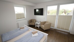 A room with two beds, a TV, a small table, and a chair. There are windows with blinds and a view of greenery outside. - Hotel Dorfkrug Büsum (Büsum)