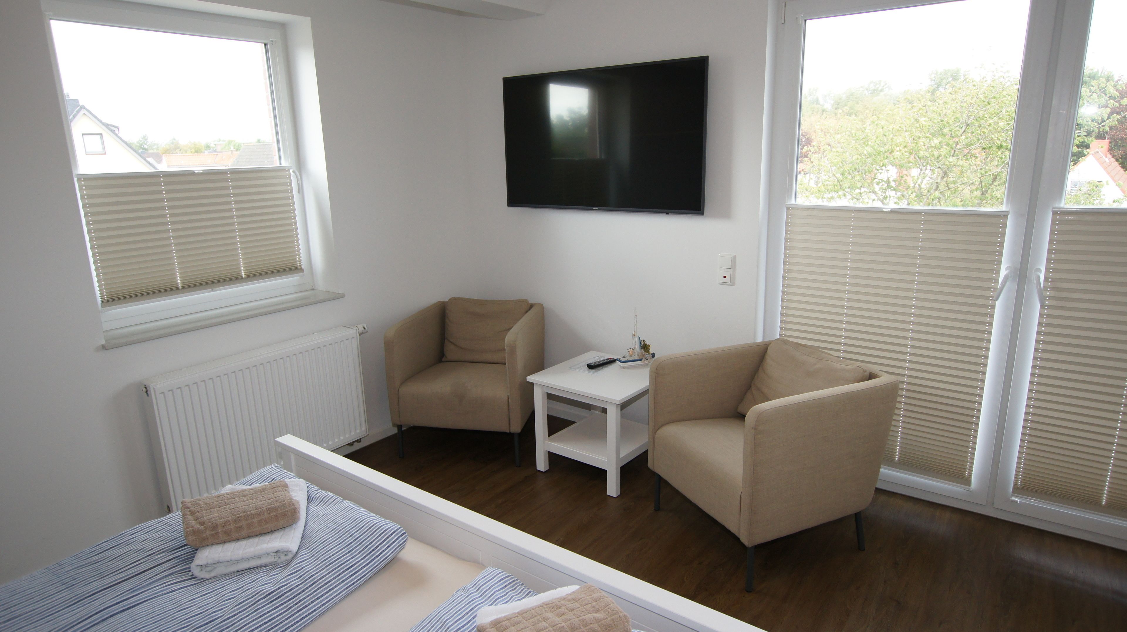 A room with two beige armchairs, a small white table, and a flat-screen TV mounted on the wall. There are windows with blinds, a radiator, and a bed with striped bedding.