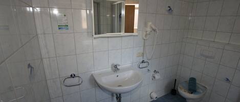 Ein Badezimmer mit weißen Fliesen, einem Waschbecken, einem Spiegel, einer Toilette und einem Handtrockner.