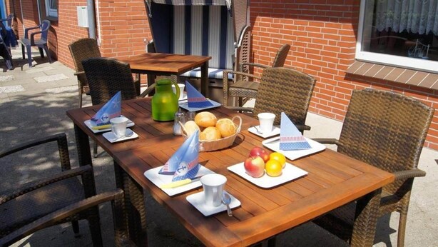 An outdoor dining area with wicker chairs and a wooden table set with plates of fruit, a pitcher, and a flag. - Hotel Dorfkrug Büsum (Büsum)