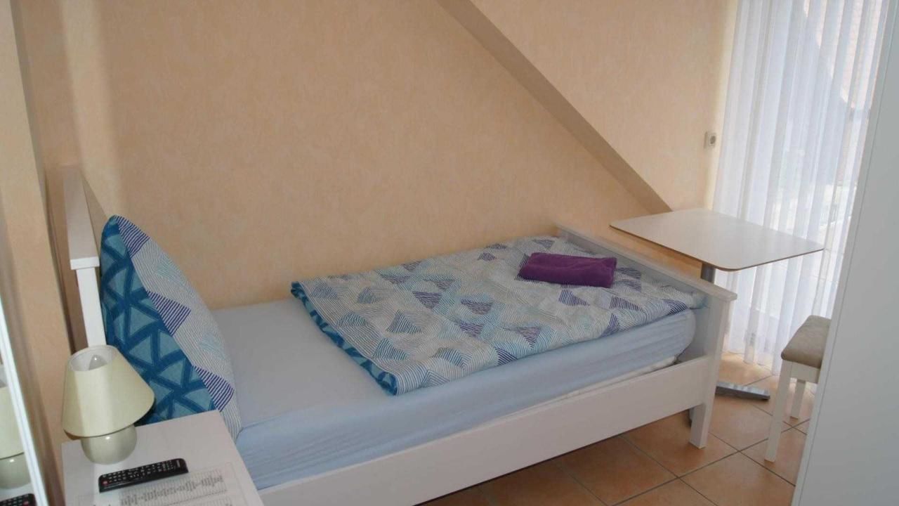 Une petite chambre avec un lit simple, un plafond en pente, une table de chevet, une lampe et une fenêtre avec des rideaux.