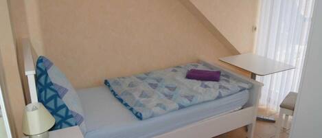 A small, single-bed room with a sloped ceiling, a bedside table, a lamp, and a window with curtains.