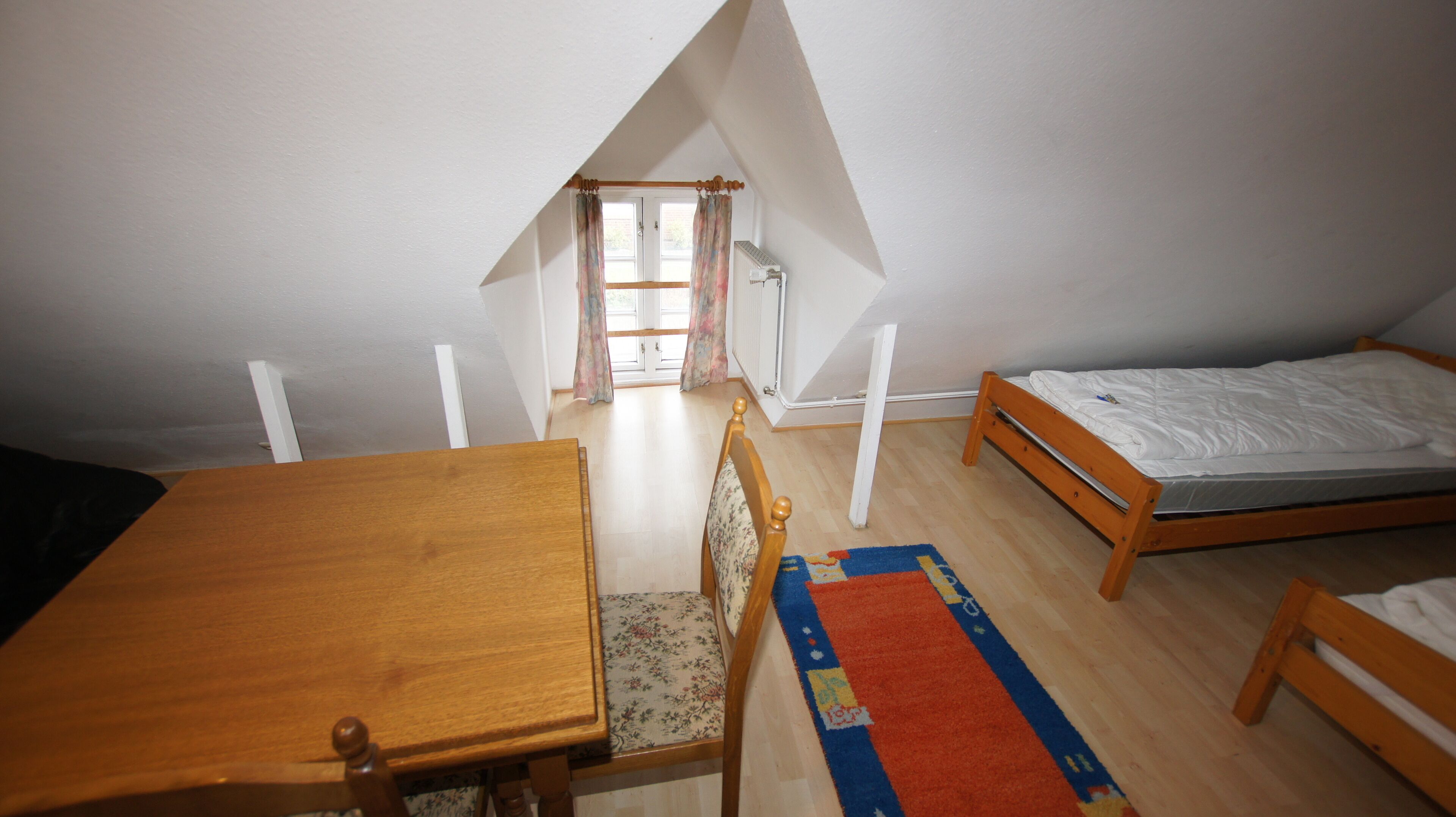 Een kleine, driehoekige slaapkamer op zolder, met een houten bed, een houten tafel en een stoel.
