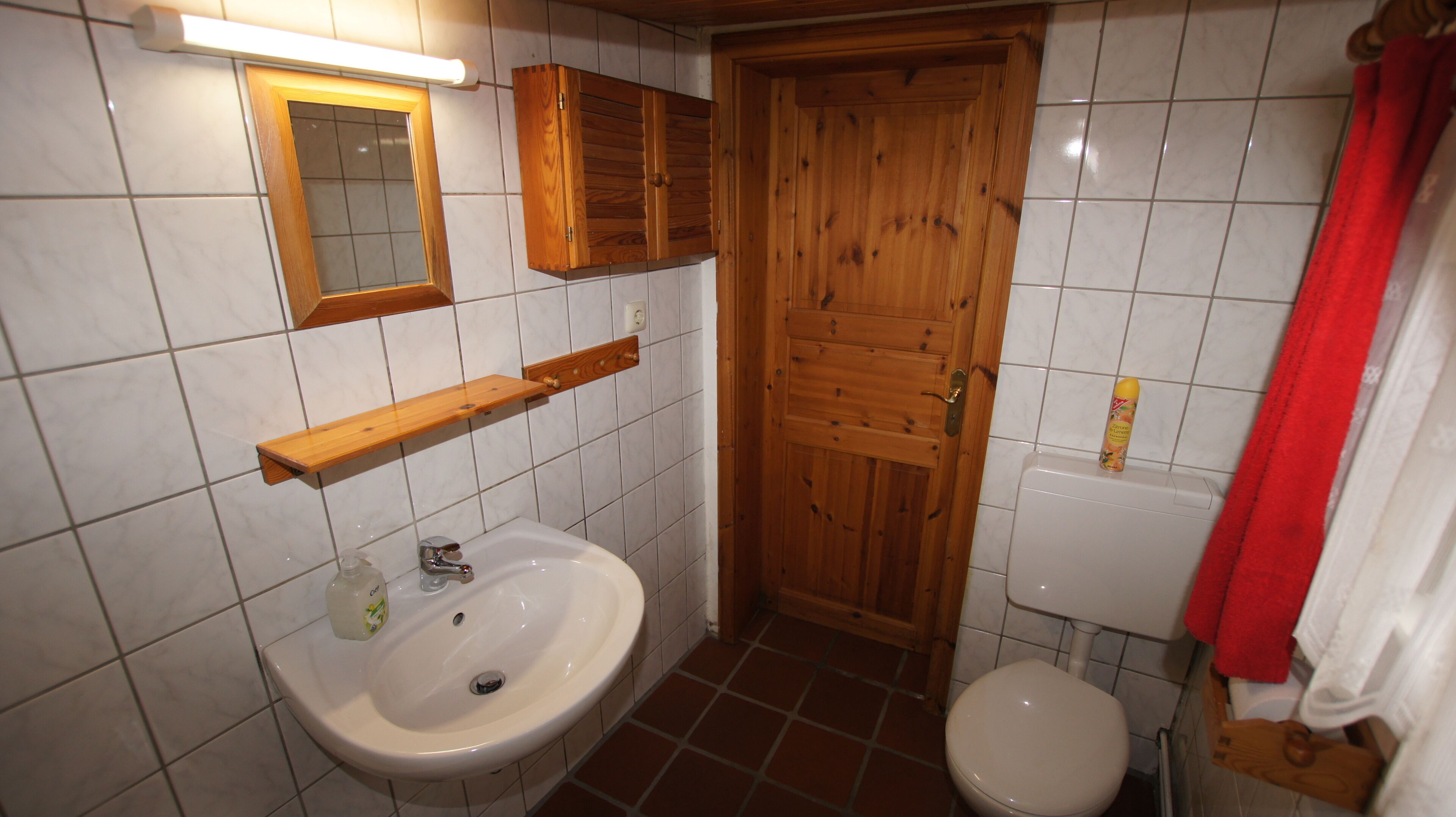 Un baño con puerta de madera, azulejos blancos, un lavabo e inodoro.