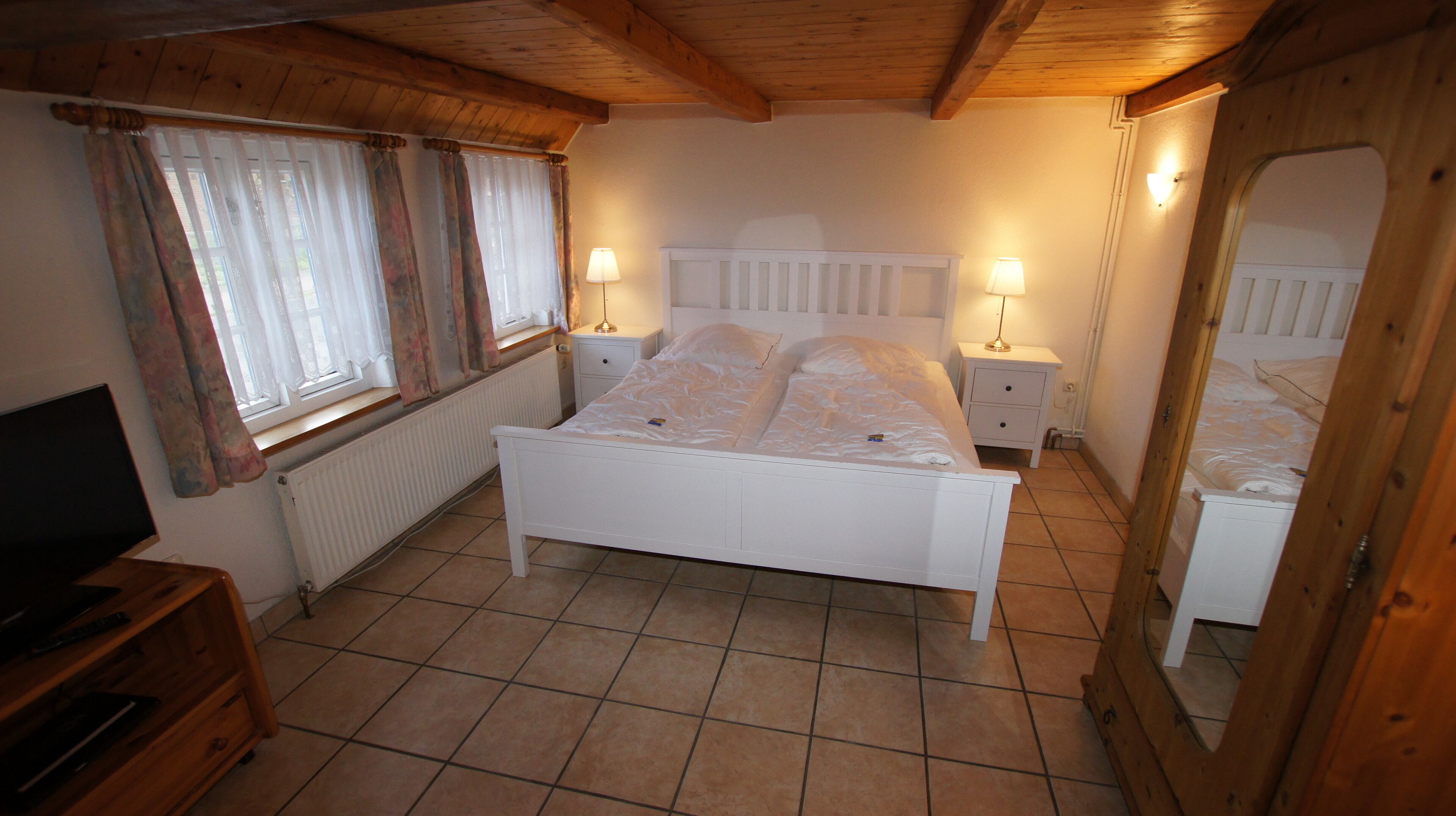 Une chambre à coucher comprenant un lit, deux tables de chevet, une commode, une coiffeuse, une fenêtre avec des rideaux et un miroir.
