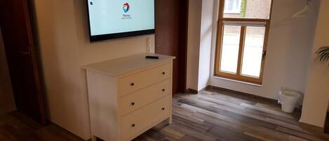 A room with a wooden floor, a white dresser, a TV mounted on the wall, and a window with blinds.