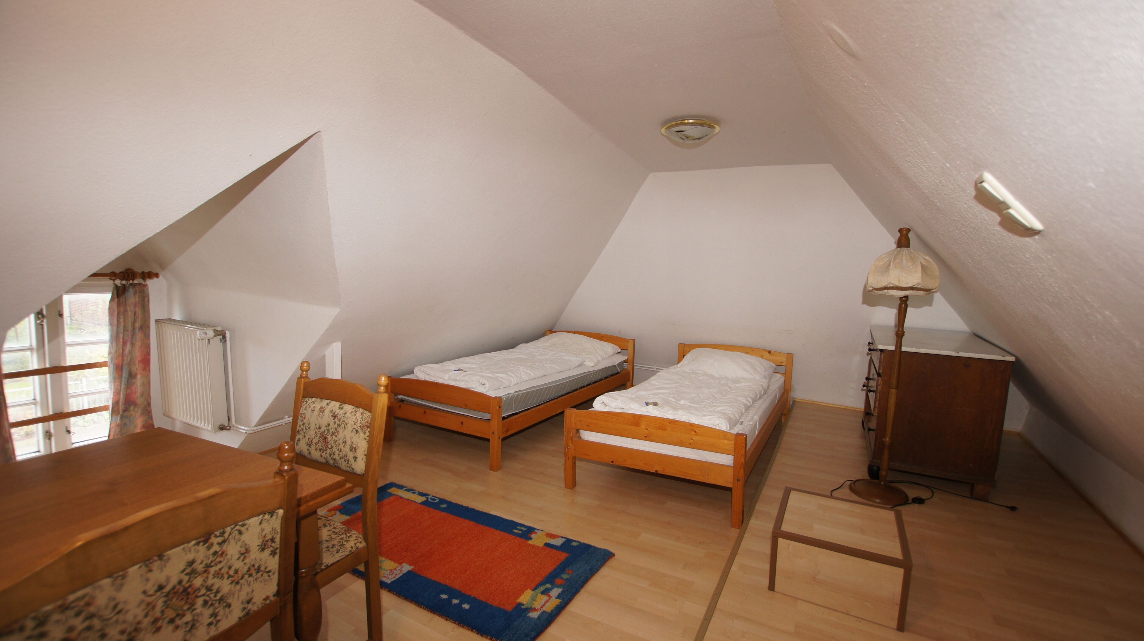 A small, attic-style room with two wooden beds, a wooden desk, a chair, a radiator, a lamp, and a window with a curtain.
