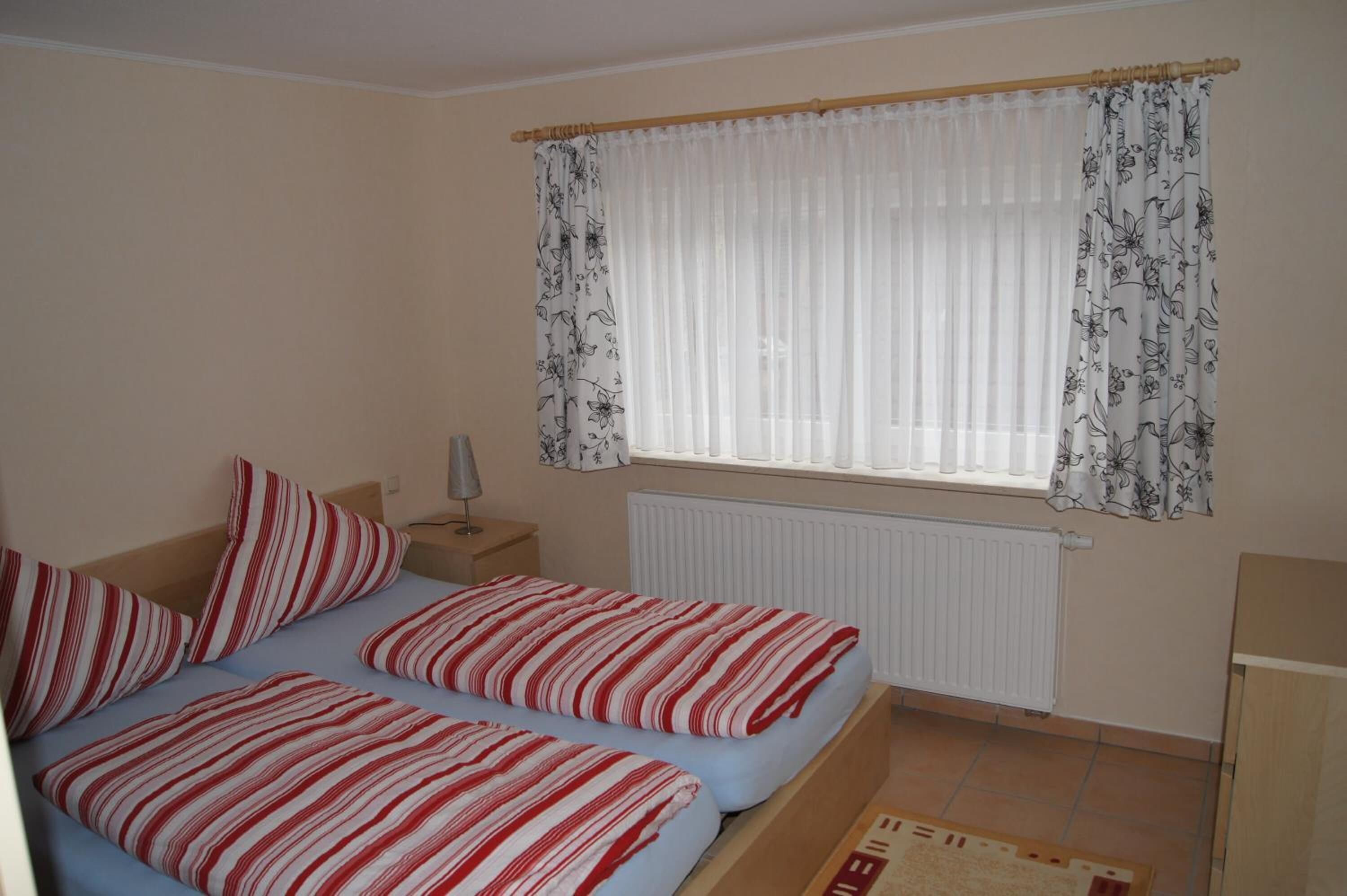 Un dormitorio con cama, dos almohadas, una mesita de noche, una lámpara, una ventana con cortinas y un radiador.