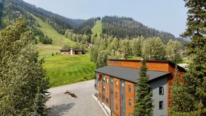 A modern building with a sign reading "The Burfield" in a mountainous area with green fields and trees. - The Burfield (Sun Peaks)