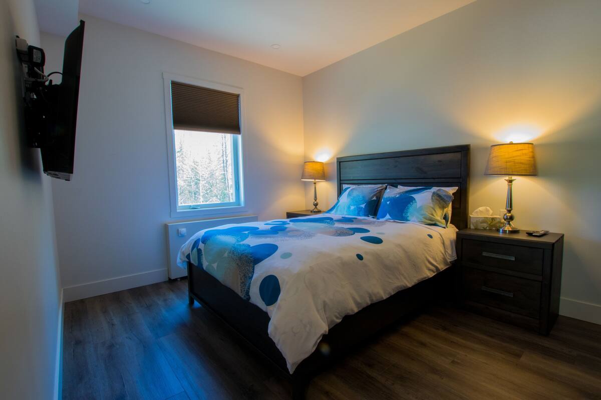 A bedroom with a bed, two bedside lamps, a nightstand, a window with blinds, and a wall-mounted TV.