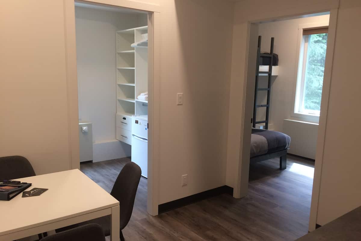 A room with a white table, chairs, a white wardrobe, a bed, and a window with a view of trees.