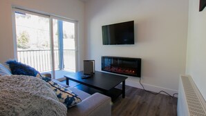A modern living room with a fireplace, a flat-screen TV mounted on the wall, and a sofa with a fluffy throw blanket. - The Burfield (Sun Peaks)