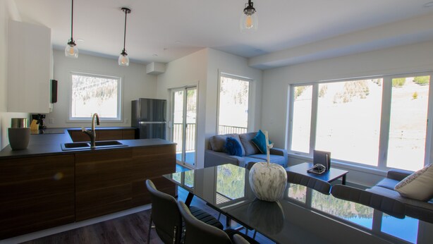 A modern kitchen with a central island, dining table, and chairs. There is a sofa with a view of the outdoors. - The Burfield (Sun Peaks)