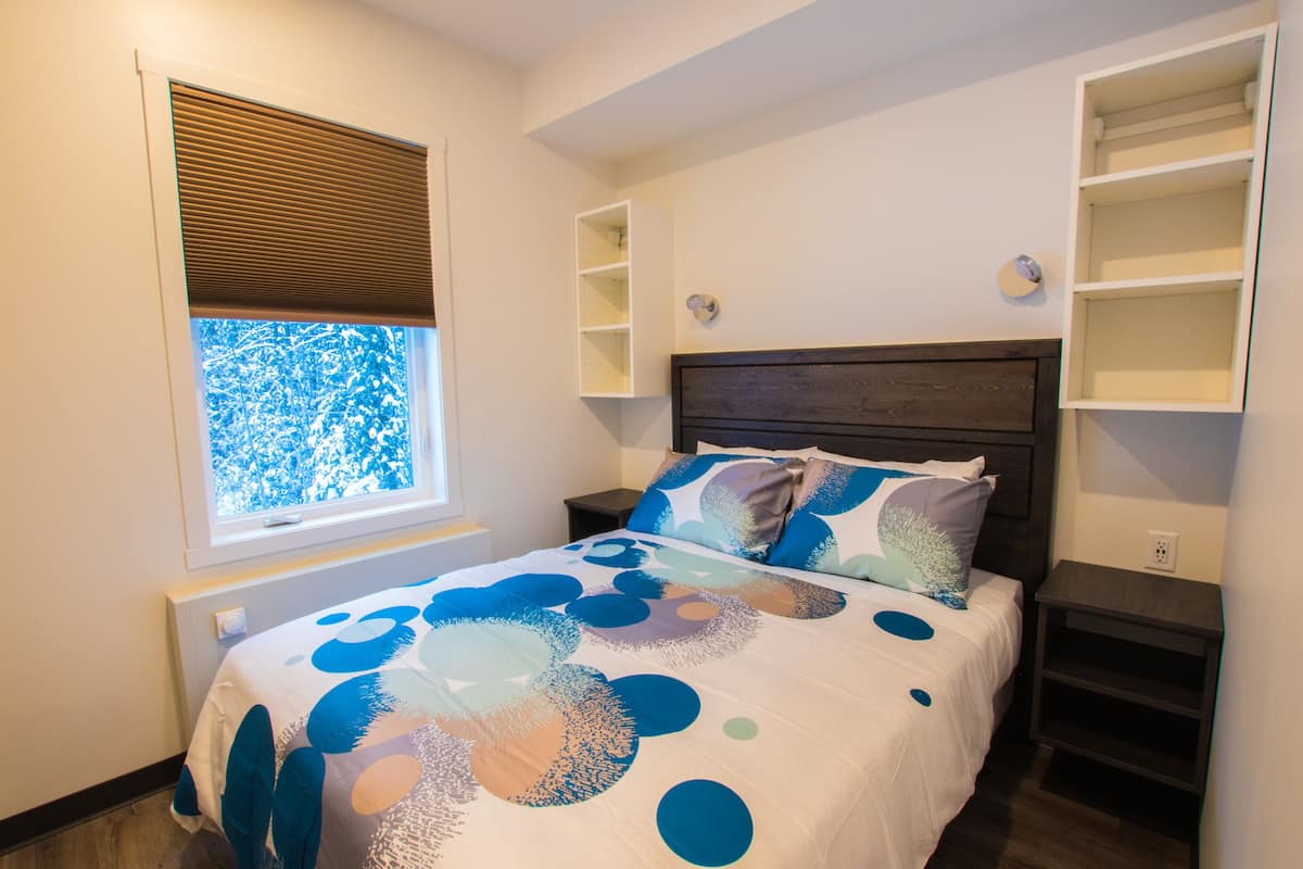 A bedroom with a bed, built-in shelving, a window with blinds, and a radiator.