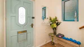 A room with a light blue door, a small mirror, a window with blue blinds, a vase with flowers, and a shelf with items. - TDM Bachelot Lettings (London)