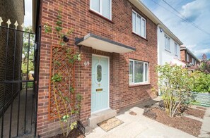 A two-story brick house with a white door and a small garden area. - TDM Bachelot Lettings (London)