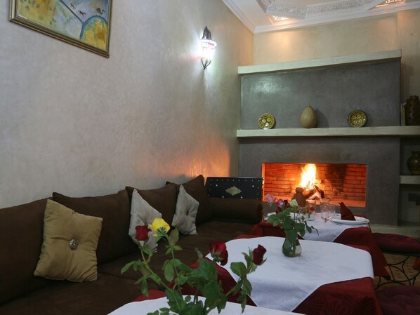 A cozy living room with a fireplace, a sofa with cushions, and a dining table set for two. - Riad El Bellar (Marrakech)