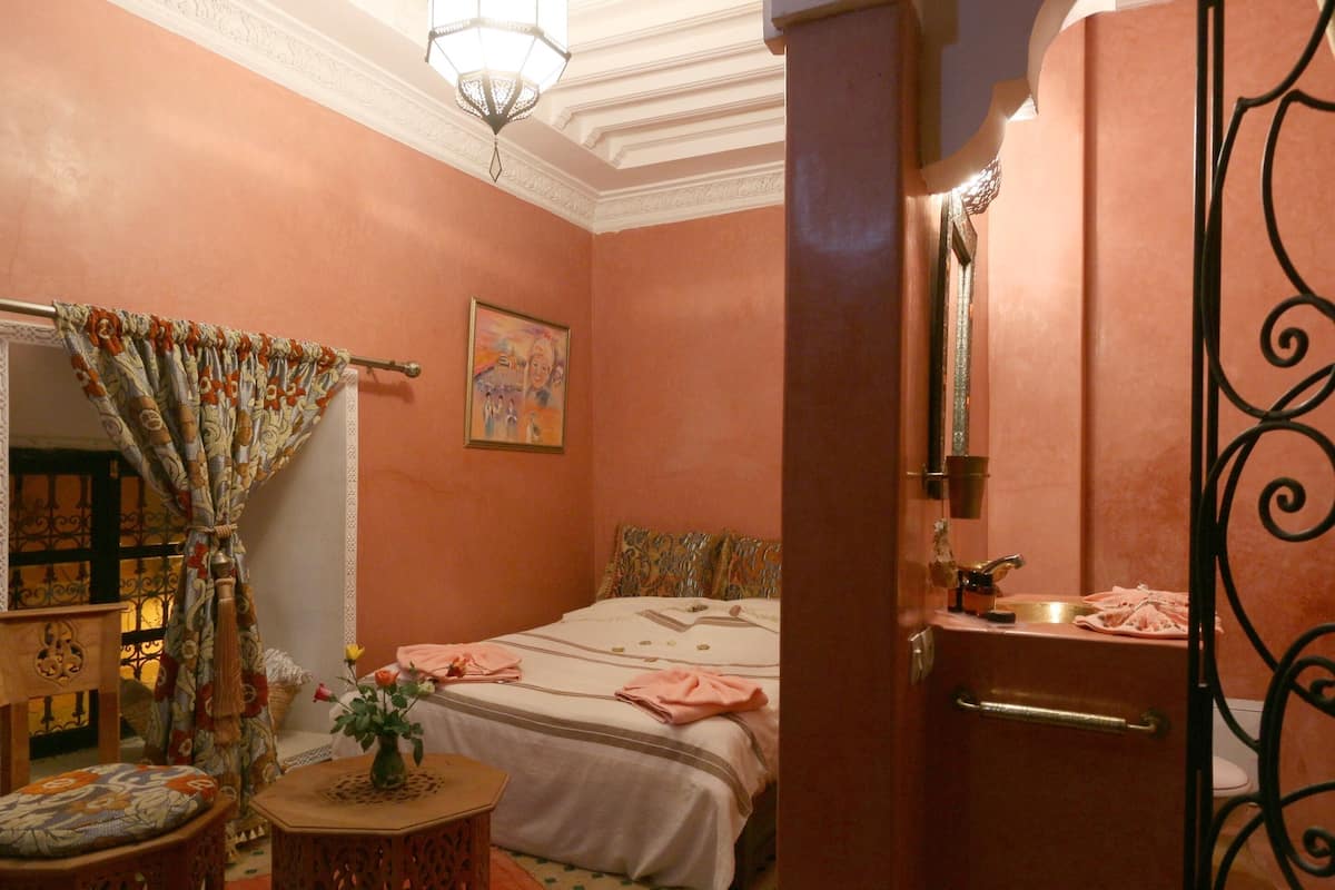A bedroom with a bed, a bedside table with a vase, a mirror, a door, and a bathroom with a sink and toilet.