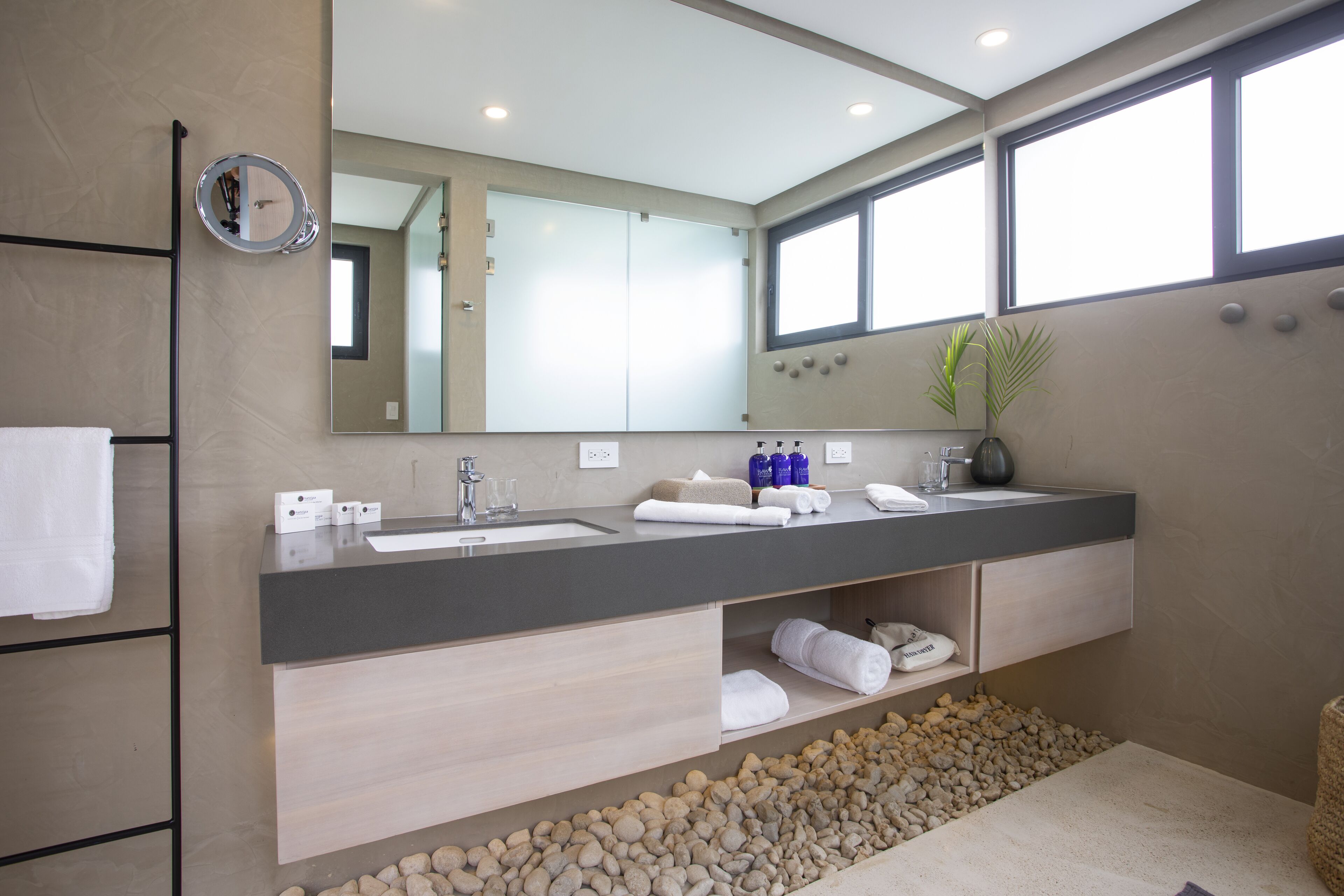 A modern bathroom with a large mirror, two sinks, a towel rack, and a plant.