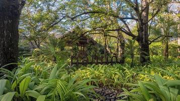 A lush garden with a wooden gazebo surrounded by dense foliage and large trees.