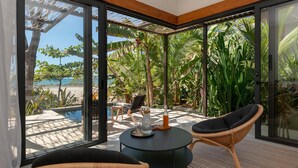 Habitación moderna con grandes puertas de vidrio que dan a una zona de piscina, con mobiliario de mimbre y una pequeña mesa con una bebida.