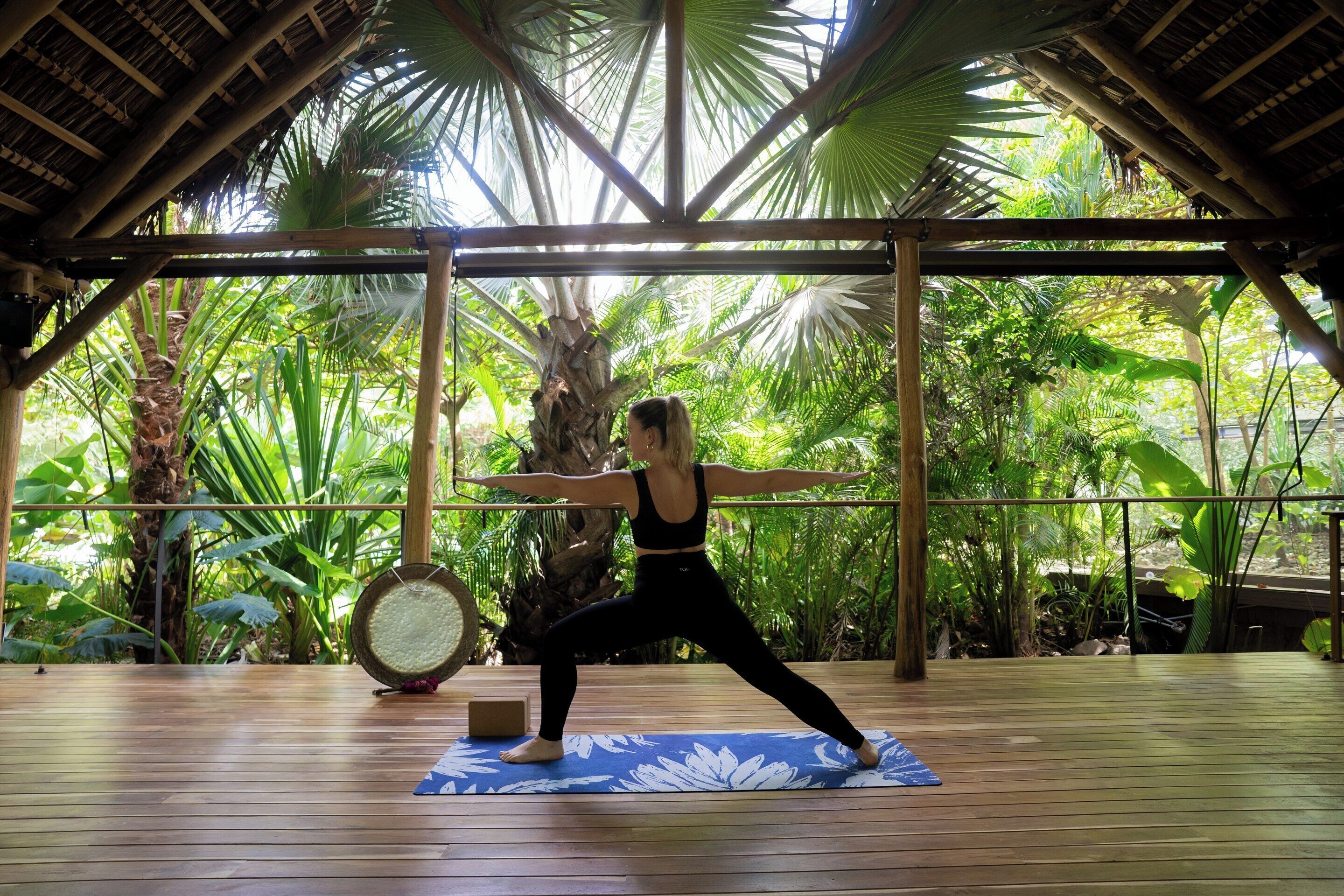 A person performing a yoga pose on a mat in a covered area with a wooden floor and tropical plants in the background.