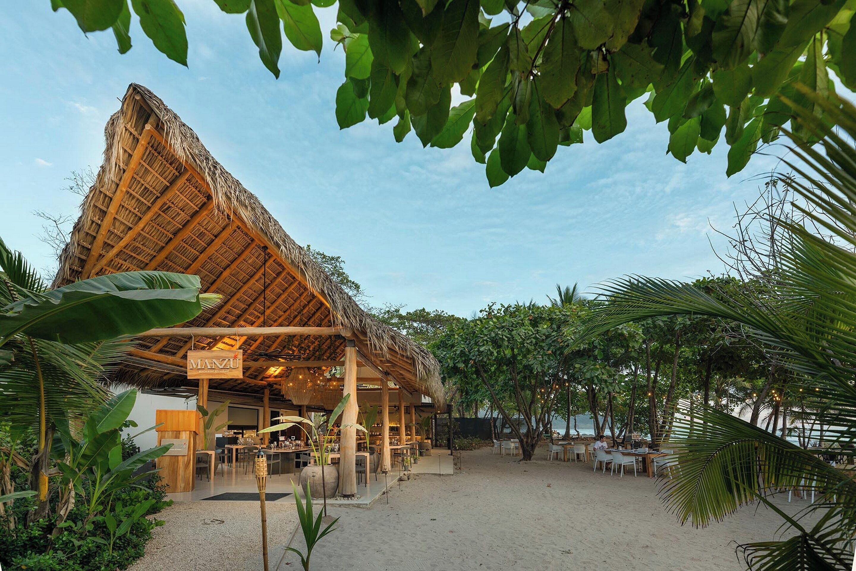 Un restaurant situé en bord de mer, doté d’un toit de chaume, de tables en plein air et d’une enseigne indiquant « MANZU ».