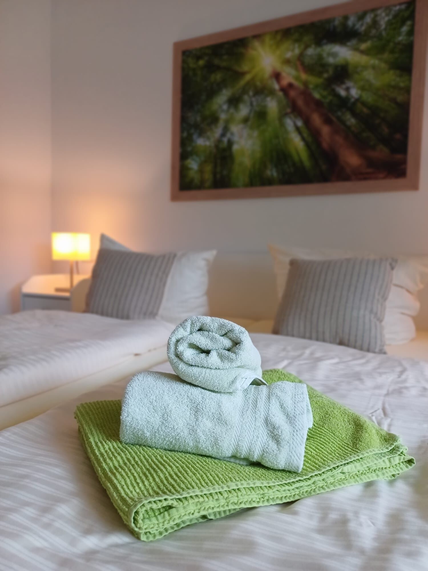 Ein ordentlich bezogenes Bett mit einem grünen und einem weißen Handtuch, ein Bild an der Wand und ein Nachttisch mit einer Lampe.