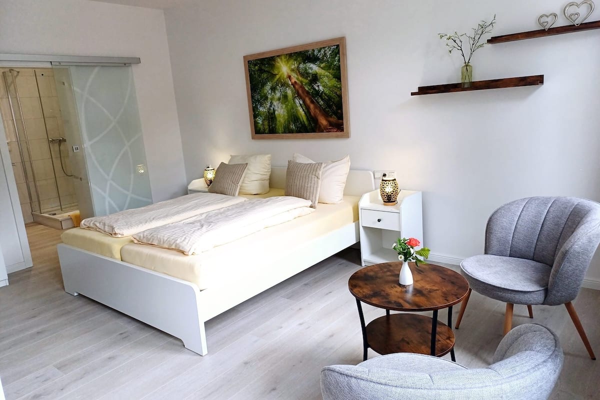 A modern bedroom with a bed, bedside tables, a chair, a small table with a vase, and a painting on the wall.
