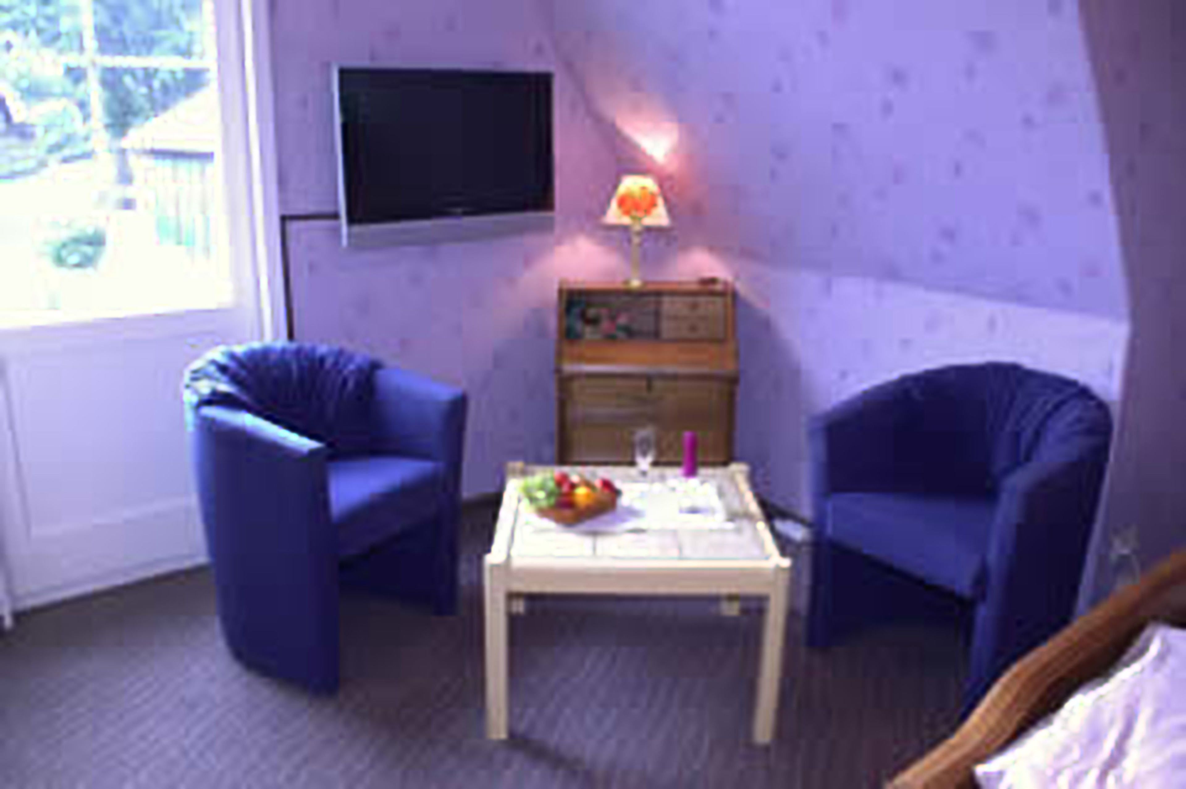 A room with two blue chairs, a small table with a fruit bowl, a lamp, and a television on the wall.