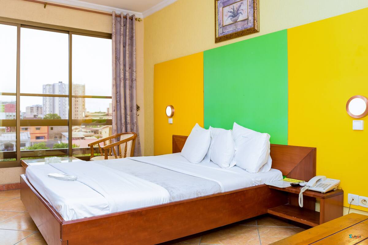 Quarto de hotel com uma cama, mesa de cabeceira com telefone e vista da cidade através de amplas janelas.