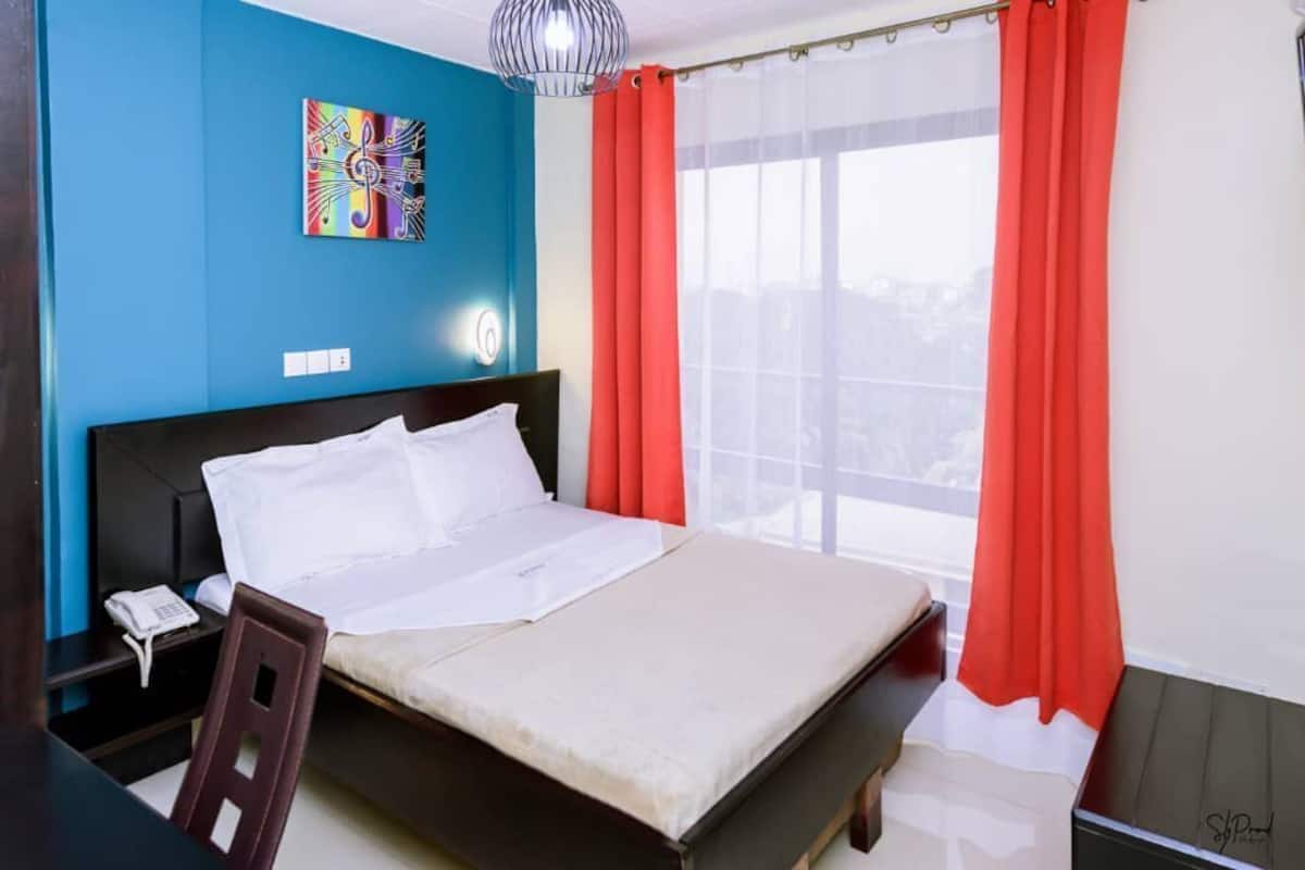 Quarto de hotel com cama, escrivaninha com telefone, cadeira, janela com cortinas vermelhas e uma obra de arte colorida na parede.