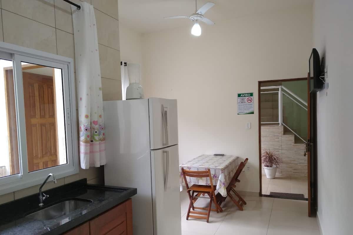 Une cuisine équipée d’un réfrigérateur, d’un évier et d’une fenêtre avec des rideaux. On y trouve une table avec des chaises et un ventilateur de plafond.