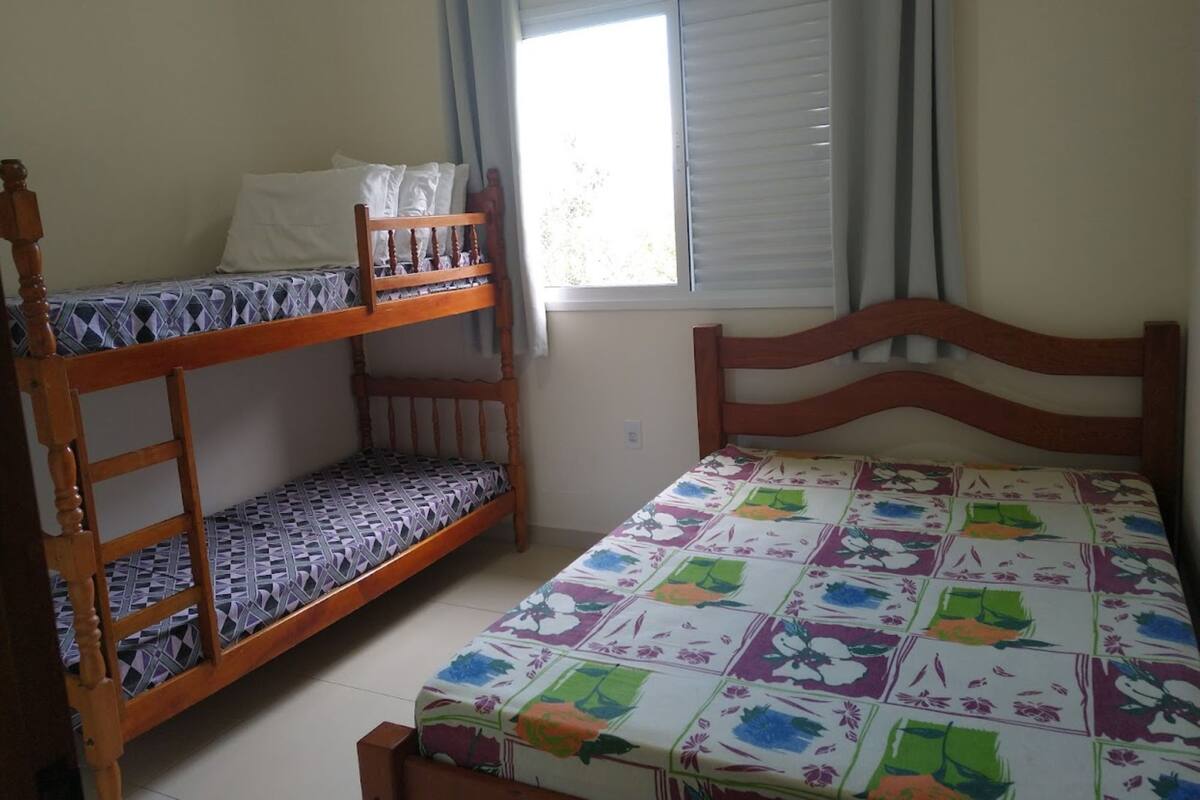 Une chambre avec des lits superposés et un lit simple, une fenêtre avec des stores et un mur uni.